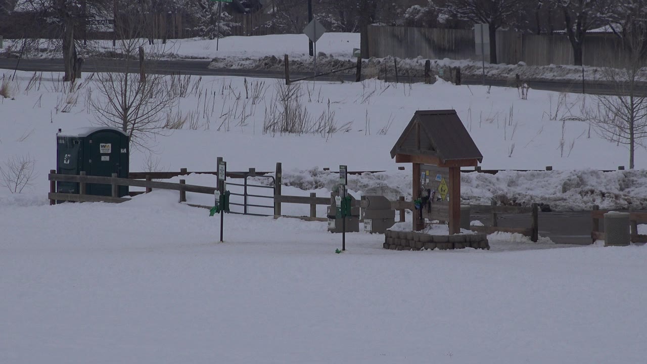 Snow-covered field with green portable toilet, fence, and roofed signboard.