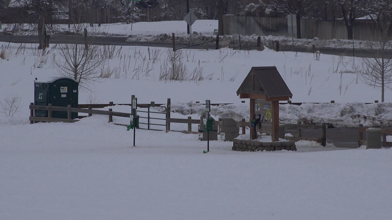 Opening frame of a snowy Arvada, Colorado outdoor area with a wooden fence, metal gate, restroom signposts, green utility boxes, bare trees, and no visible people.