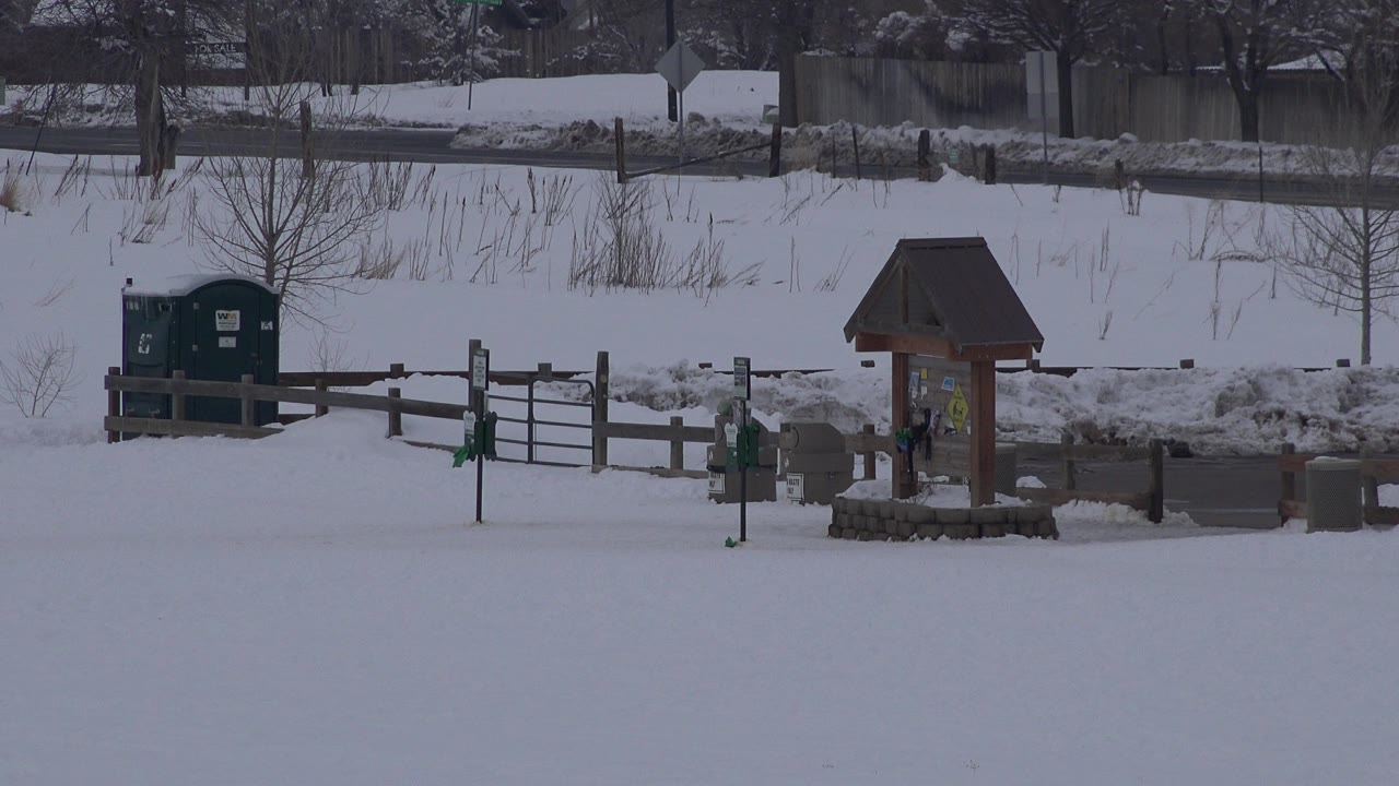 Opening frame of an Arvada, Colorado snow-covered public area with a low fence, green bins, a wooden information kiosk, bare trees, and no visible people.