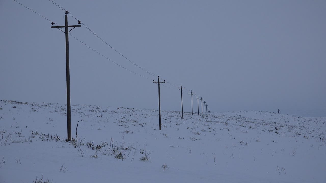 At 0:00 in Arvada, Colorado, a tall foreground telephone pole stands left of center while smaller poles recede across a snow-covered field under a blue-gray sky, with no visible people.