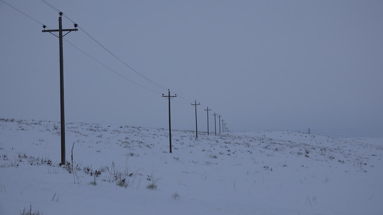 At 0:00 in Arvada, Colorado, a tall telephone pole stands at left with smaller poles receding across a snow-covered field under blue-gray overcast light, with no visible people.