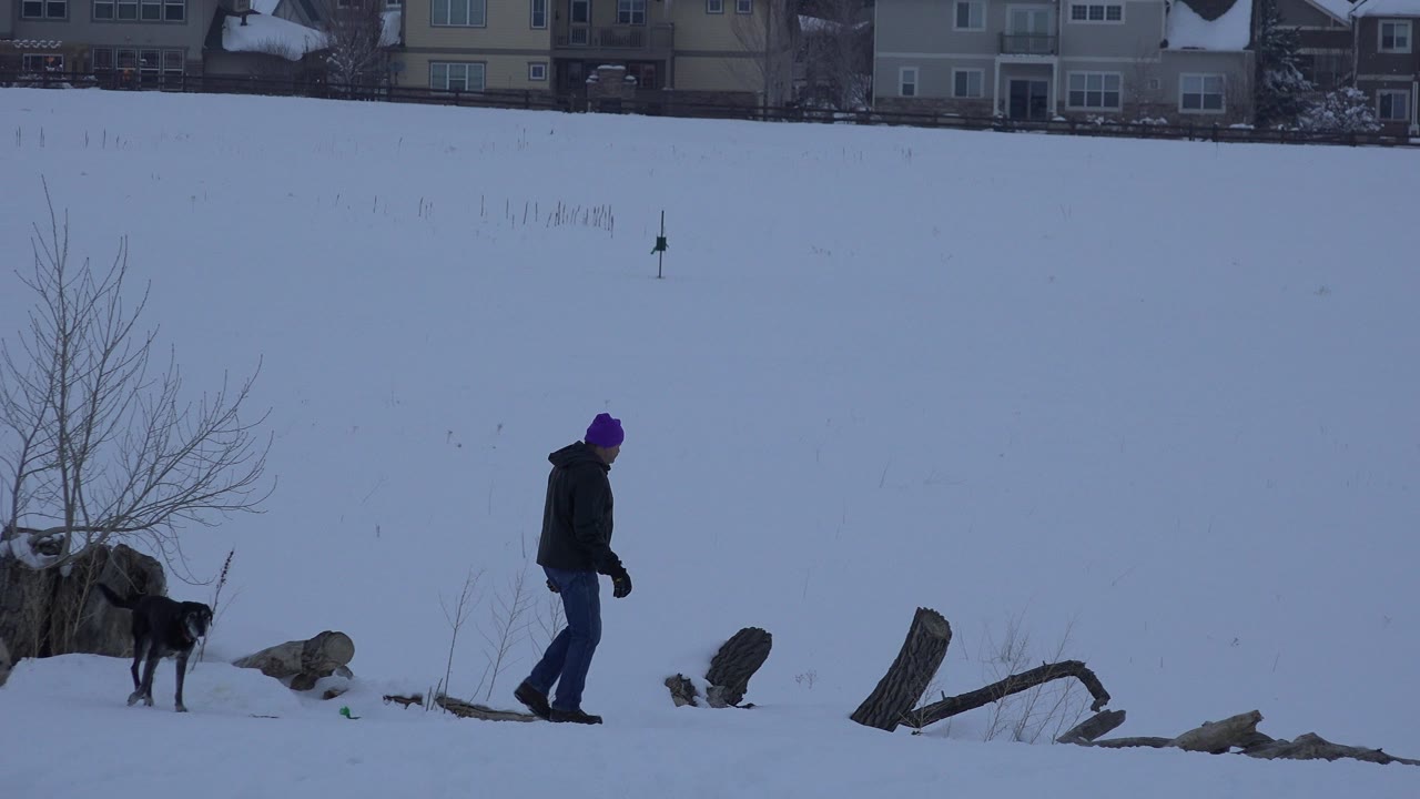 Man in a purple hat and black dog in a snowy open area.