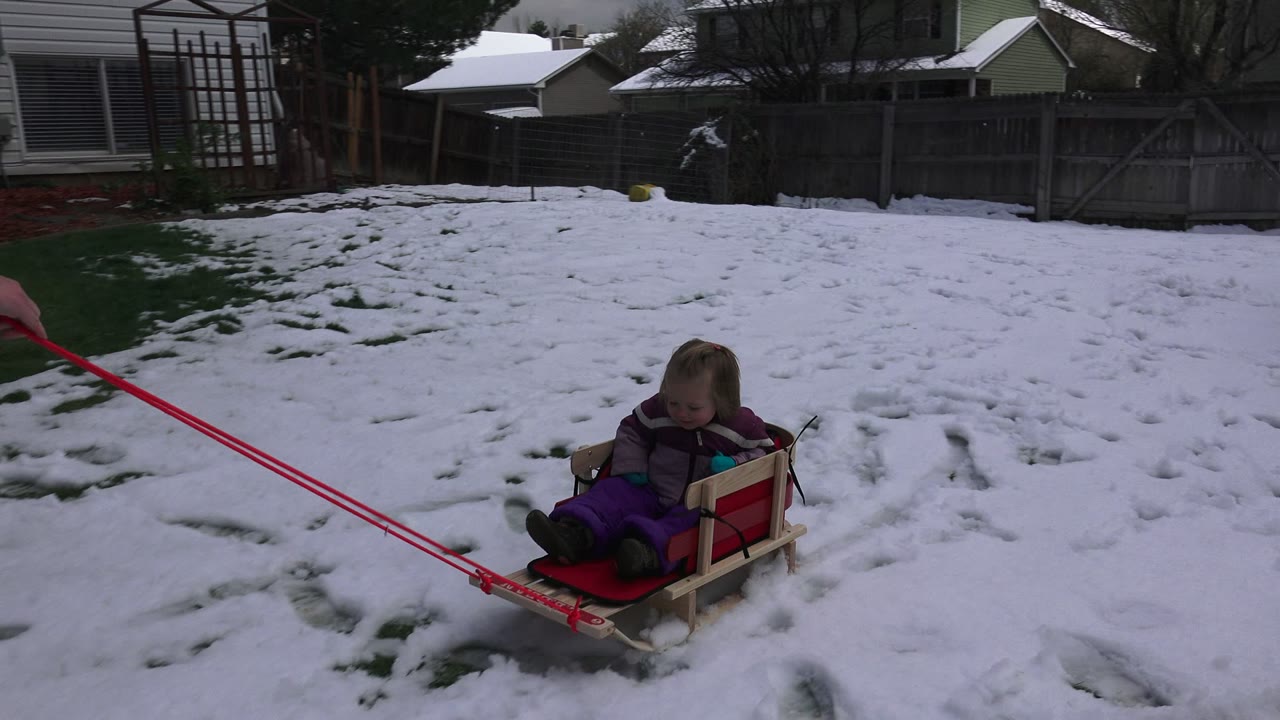 Toddler sitting on a wooden sled in a snowy backyard.
