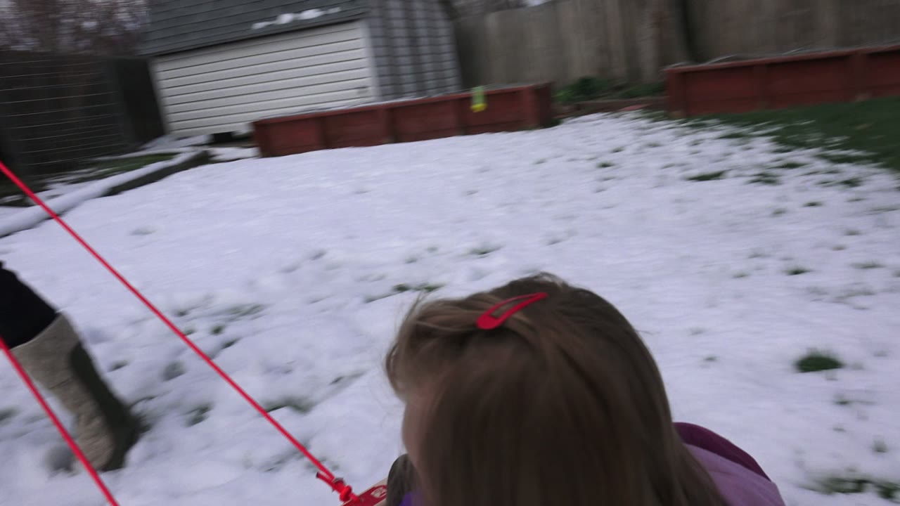 Child seen from behind in a sled on snowy grass while an adult walks nearby.