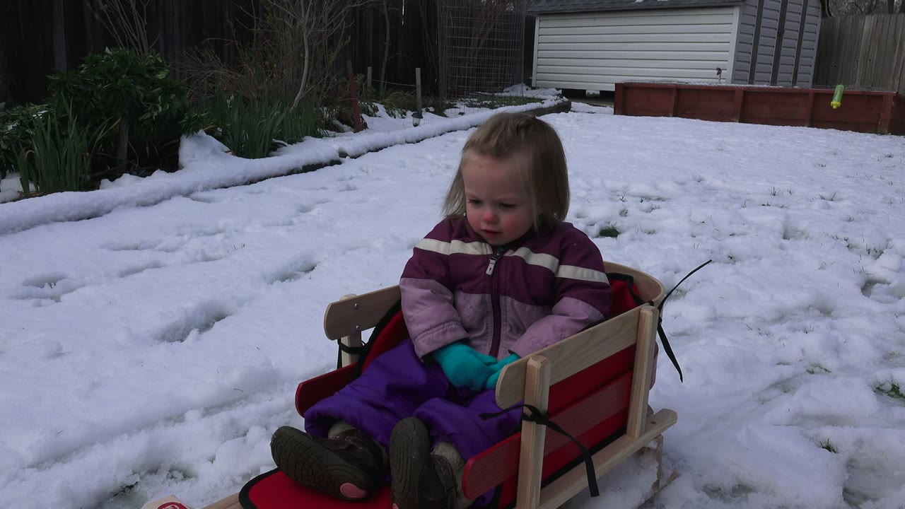 At 0:01, the child remains seated in the red sled, looking downward while her teal mittens rest together against her purple snow clothing.