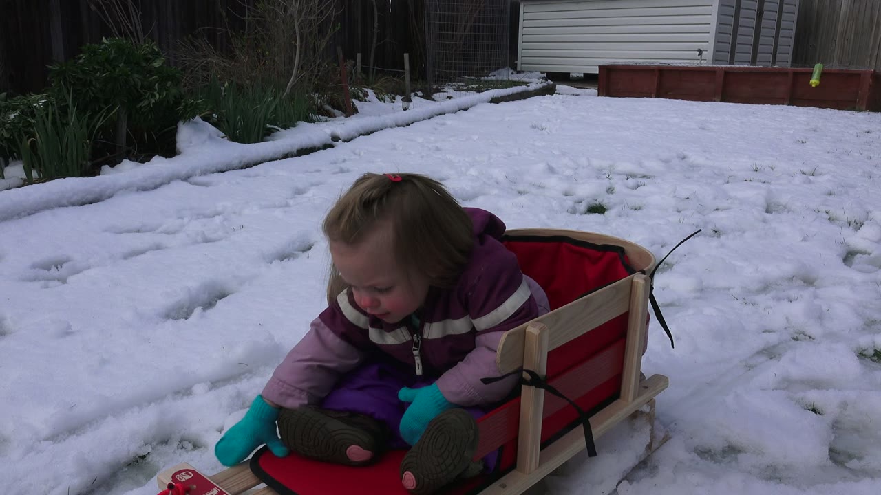 Young girl leaning forward in a red sled on snowy ground.
