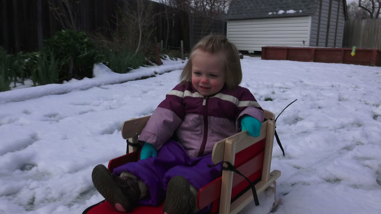 Toddler seated in a red sled on snowy backyard ground.