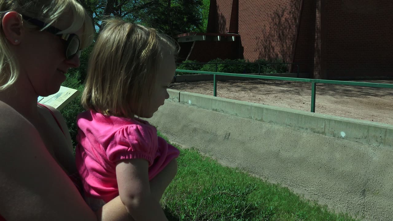 In Westminster, Colorado, a woman in dark sunglasses holds a young girl in a pink shirt beside a concrete wall, green railing, grass, trees, and red brick structures.
