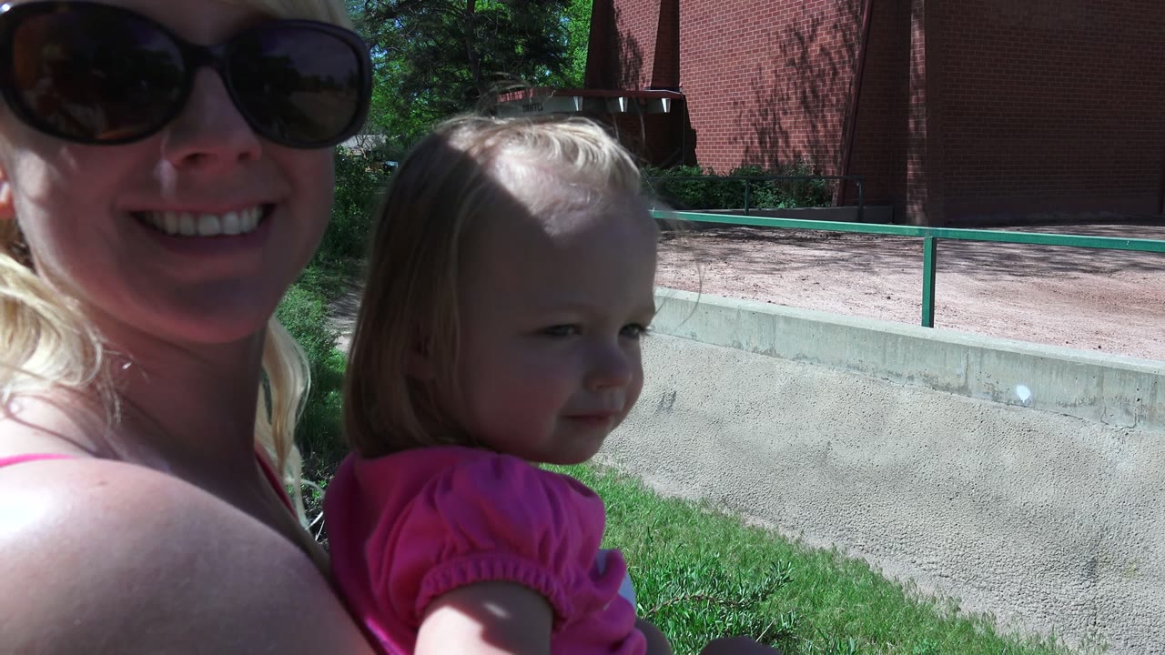 In Westminster, Colorado, a blonde woman in dark sunglasses smiles at the camera while holding a young blonde child in a pink shirt near grass, a low wall, green railing, and a brick building.