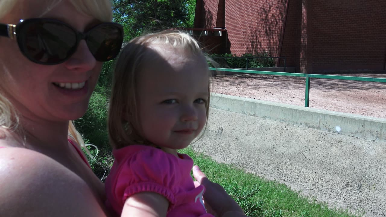 In Westminster, Colorado, the woman turns slightly toward the child while smiling, with the child's face more centered against the green rail, grass, and red brick background.