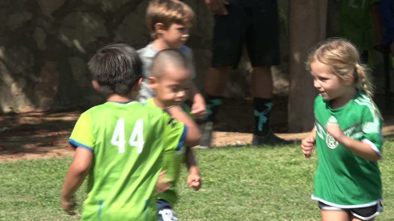 Several children in green soccer uniforms run across the grass in San Jose del Cabo, Mexico, with one player facing away in a number 44 shirt.