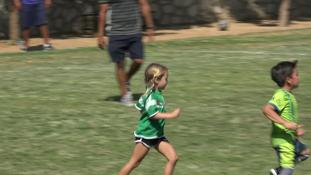 Children in green soccer jerseys run on a grass field in San Jose del Cabo, Mexico, with an adult and stone wall visible behind them.