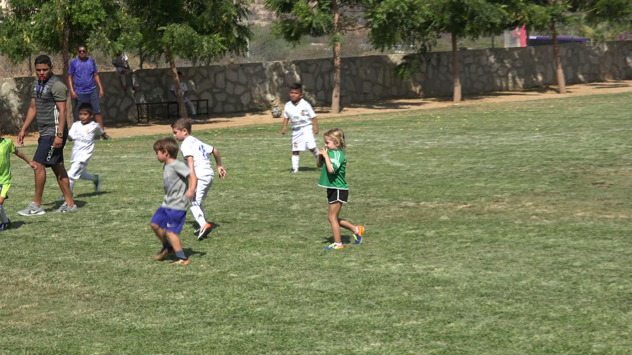 Children playing soccer on a grassy field with adults nearby.