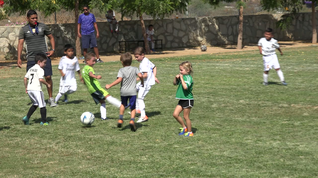 Several children converge on a soccer ball in San Jose del Cabo, Mexico, with players in white and green uniforms and a stone wall behind the field.