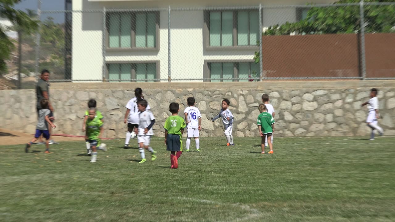 Children in green and white soccer uniforms move toward the corner area in San Jose del Cabo, Mexico, with an adult trainer standing near the play and a stone wall behind them.