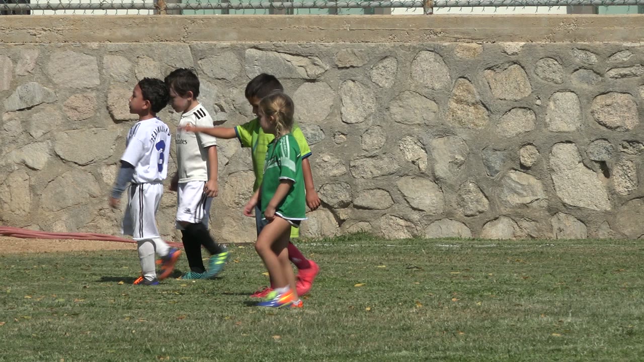 A group of young players in white and green uniforms walks left to right on the grass in San Jose del Cabo, Mexico, with the stone wall filling the background.