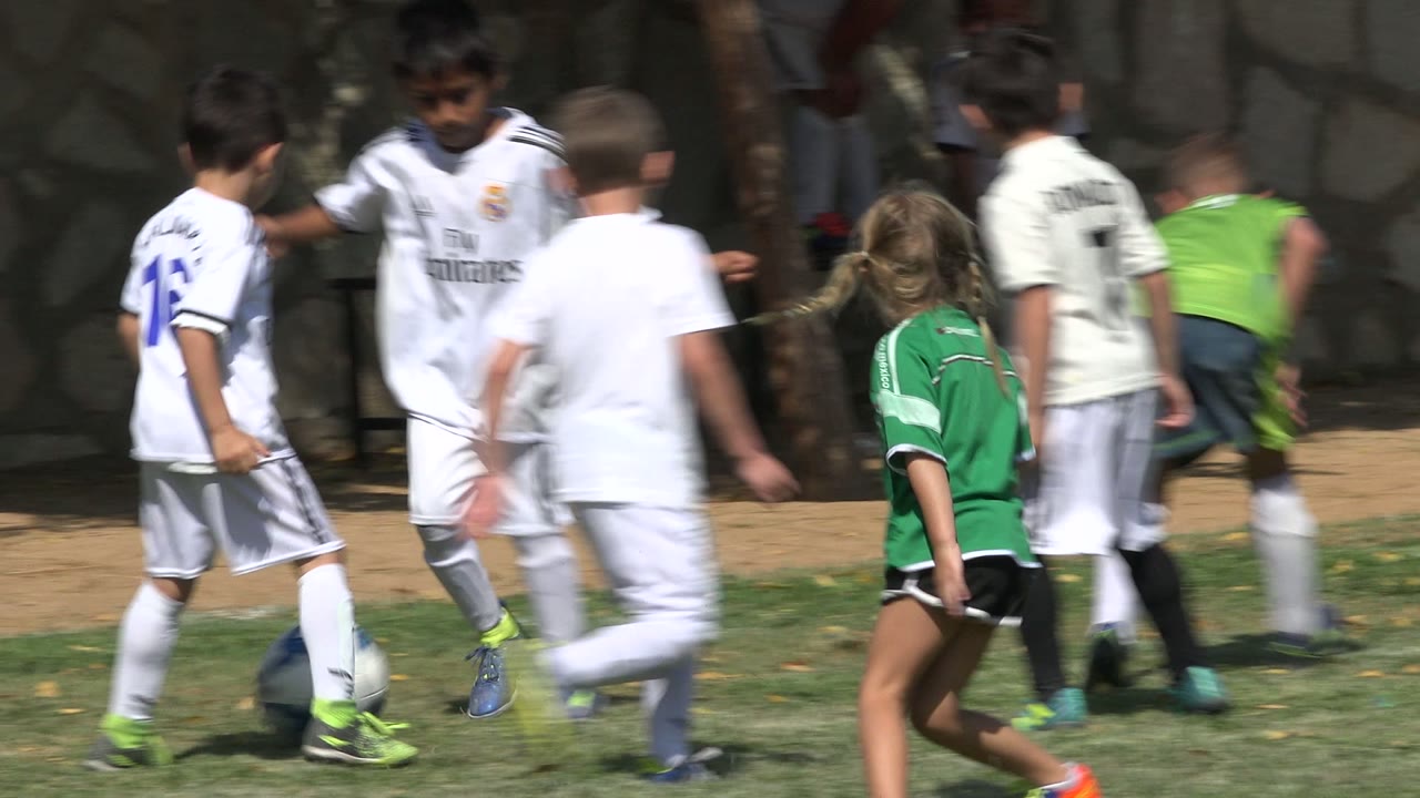 Boys in white soccer kits and a girl in a green jersey walk and run near a stone wall in San Jose del Cabo, Mexico, with strong sunlight on the field.