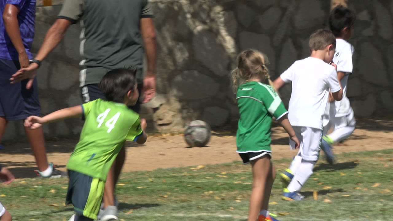 Children in green and white soccer uniforms move near a ball on a grass field in San Jose del Cabo, Mexico, with adult legs partly visible at the left edge.