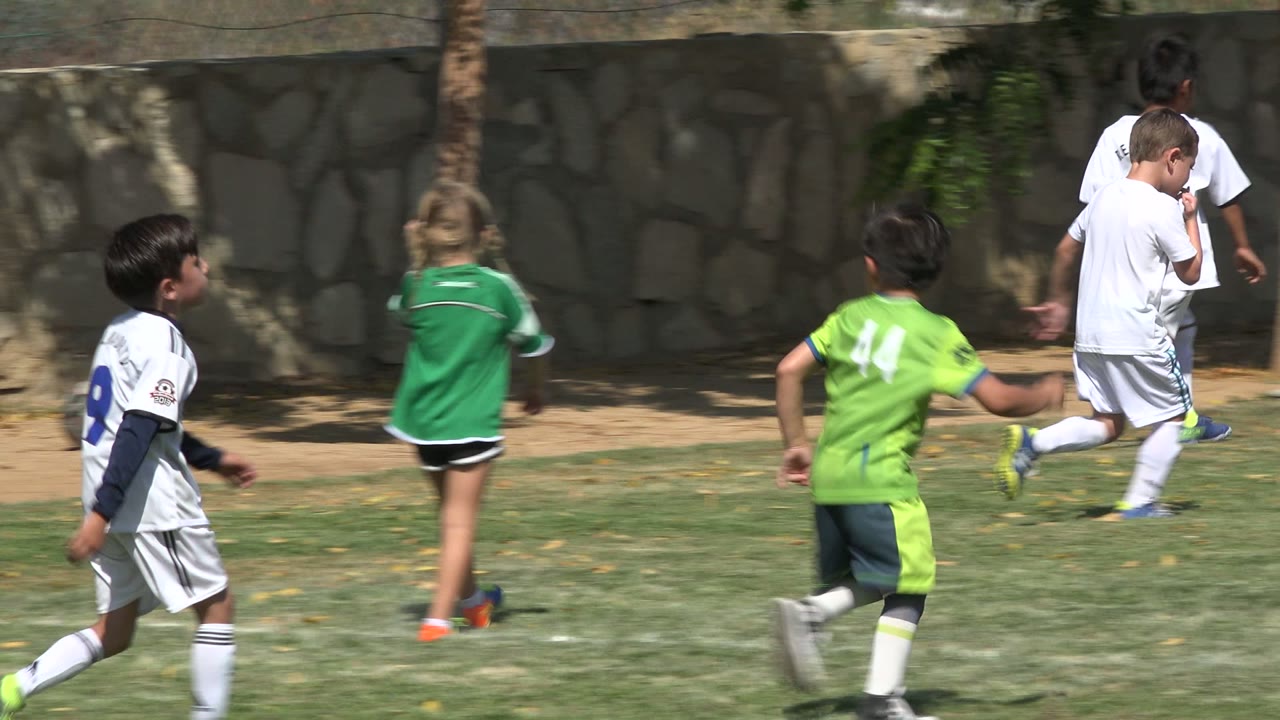 Children in white and green soccer uniforms run across a sunlit grass field in San Jose del Cabo, Mexico, with trees and a stone wall behind them.