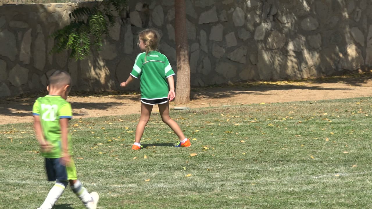 In San Jose del Cabo, Mexico, a girl in a green soccer shirt and orange shoes walks across a sunlit grass field while a boy in green moves through the foreground.
