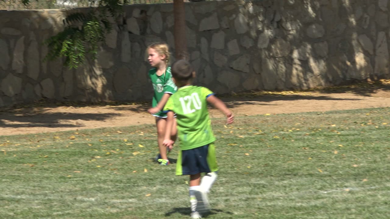 In San Jose del Cabo, Mexico, the girl in green stands near a boy in a bright green number 120 jersey on the grass with a shaded stone wall behind them.
