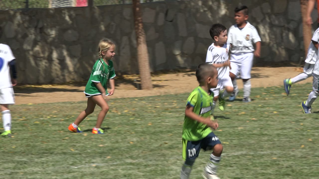 Children in green and white soccer uniforms run across a grass field in San Jose Del Cabo, Mexico, with a stone wall and palm trunks behind them.