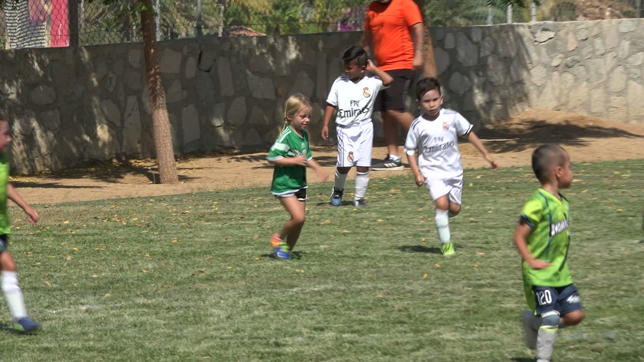 Young soccer players in green and white kits run on the field in San Jose Del Cabo, Mexico, while an adult in an orange shirt stands near the stone wall.