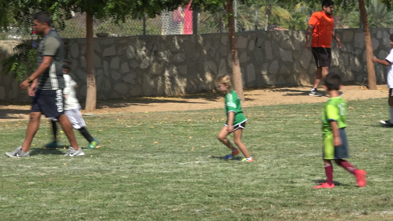 A coach and several children in soccer kits cross the grass field in San Jose del Cabo, Mexico, with trees shading the stone wall in the background.