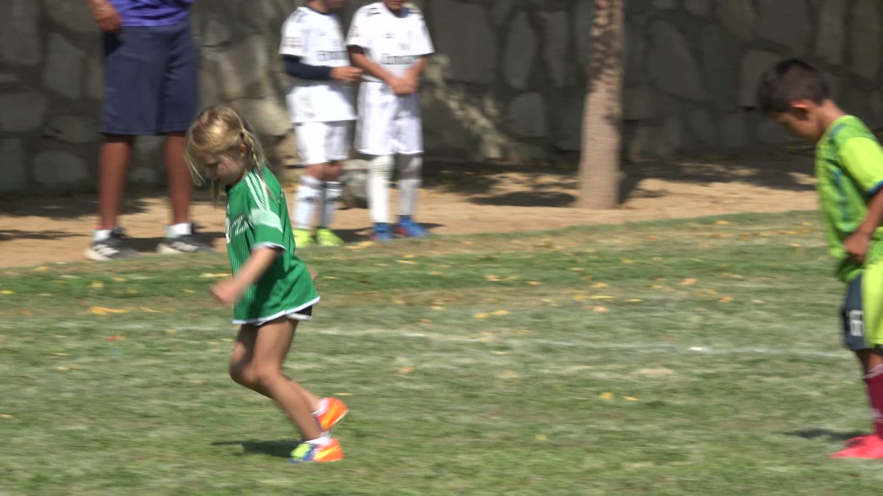 Children in green and white soccer uniforms run across a sunny grass field in San Jose del Cabo, Mexico, with adults and a stone wall behind them.