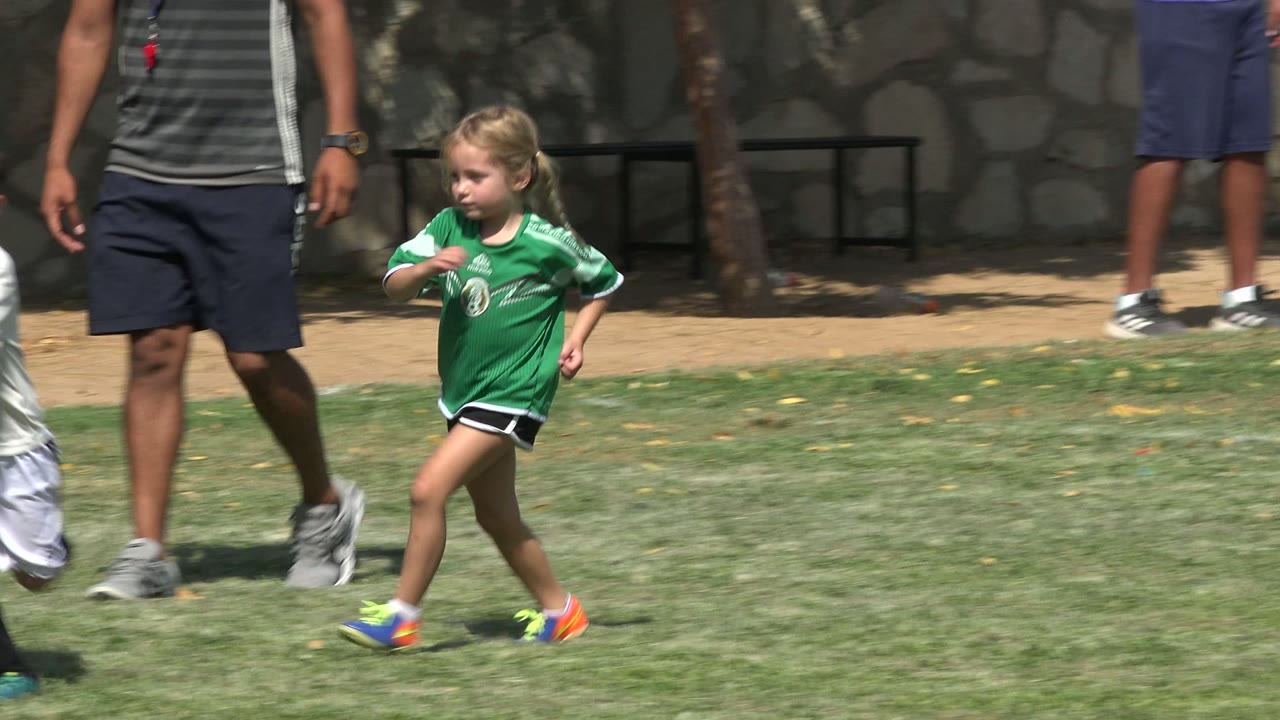 A blonde child in a green soccer jersey runs on the grass in San Jose del Cabo, Mexico, while an adult supervisor stands near the sideline and stone wall.