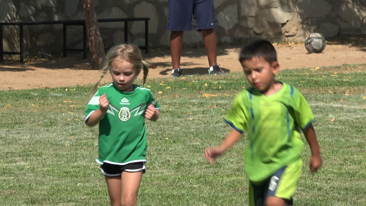 Young girl in a green soccer jersey on a grass field with other players nearby.
