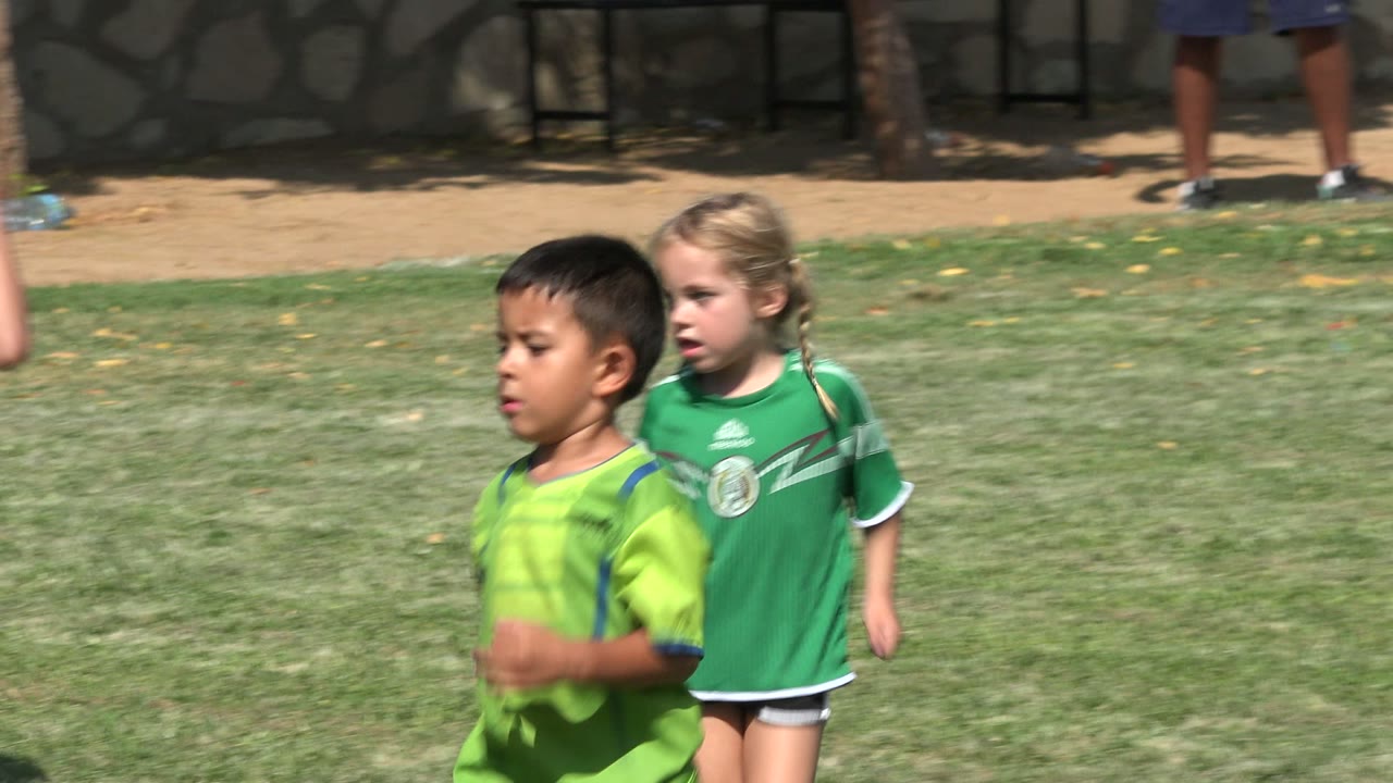 A child in a lime green soccer shirt runs through the midground on a grass field in San Jose del Cabo, Mexico, with a soccer ball and sideline figures behind him.