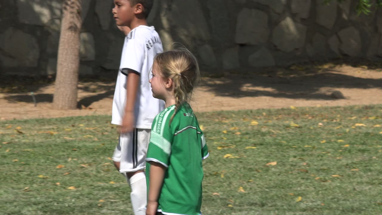 Children in green and white soccer jerseys stand and move on a sunlit grass field in San Jose del Cabo, Mexico, with a stone wall and tree trunk behind them.