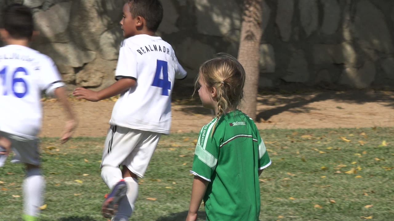 Young players in white and green jerseys run across the grass in San Jose del Cabo, Mexico, with motion blur and a shaded stone wall in the background.