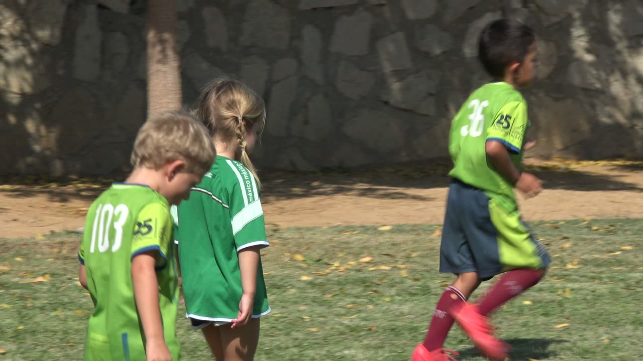Children in green jerseys walk beside a soccer ball.