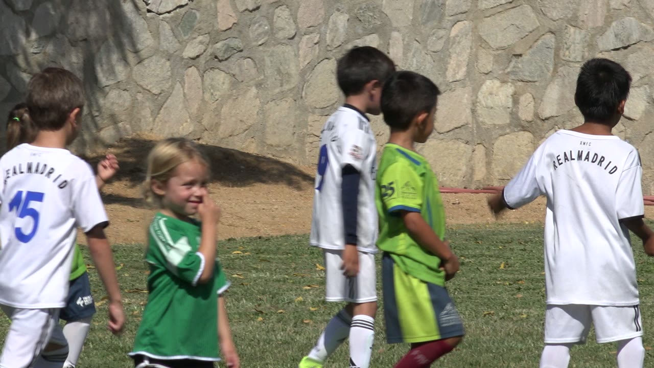 Several children in white and bright green soccer uniforms walk through the grass in San Jose Del Cabo, Mexico, with jersey numbers visible and a stone wall behind them.