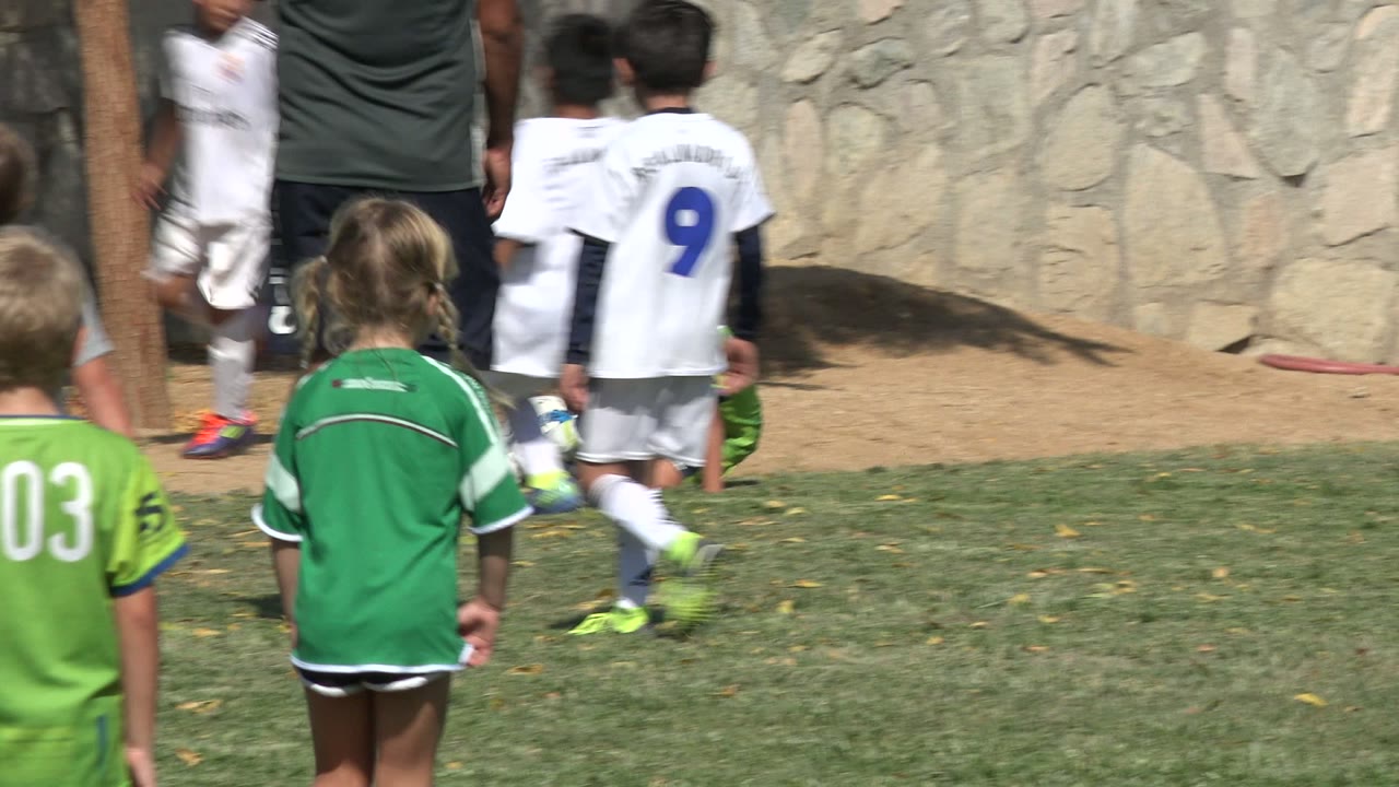 Children in white and green soccer uniforms move across the grass in San Jose del Cabo, Mexico, with a soccer ball rolling near the center and a stone wall in back.
