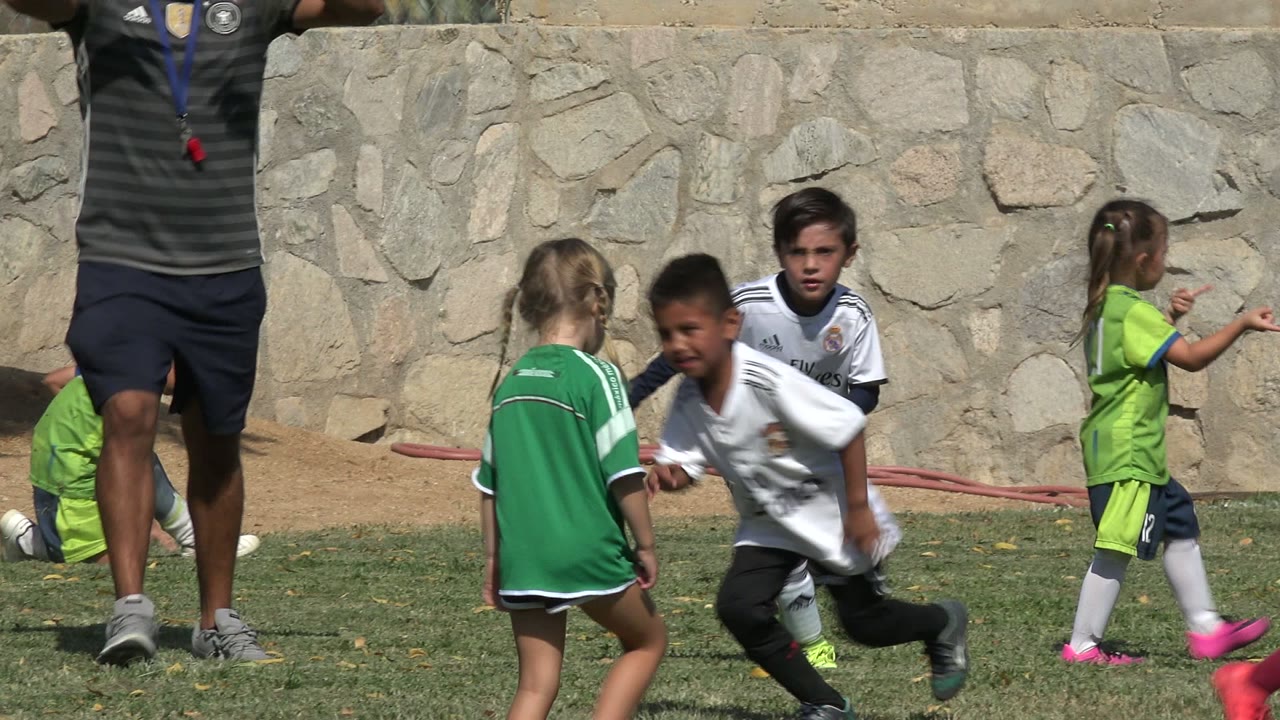 Children in green and white soccer uniforms move across the grass in San Jose del Cabo, Mexico, while an adult stands partly cropped near the center background.