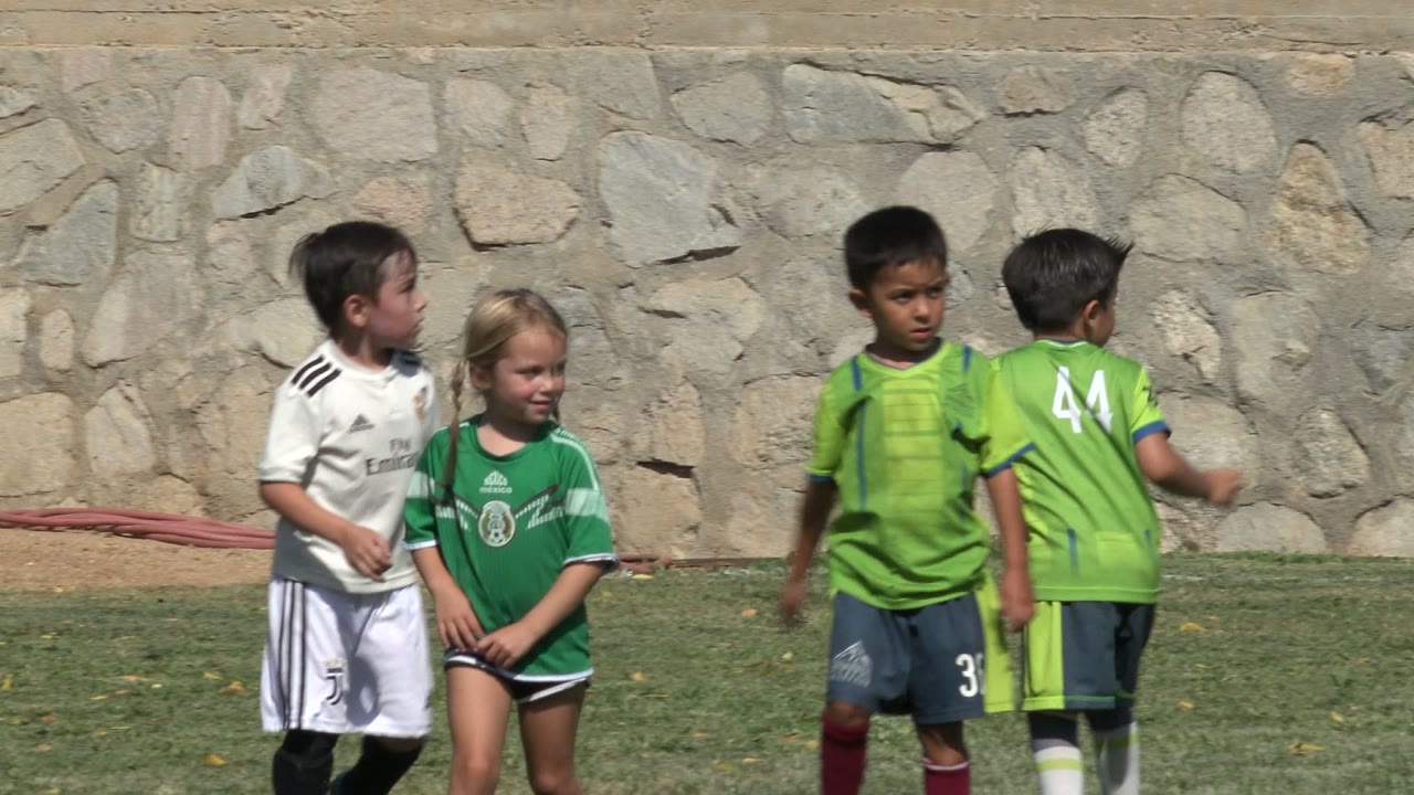 Children in green and white soccer uniforms stand on a grass field in San Jose Del Cabo, Mexico, with a stone wall behind them at the start of the clip.