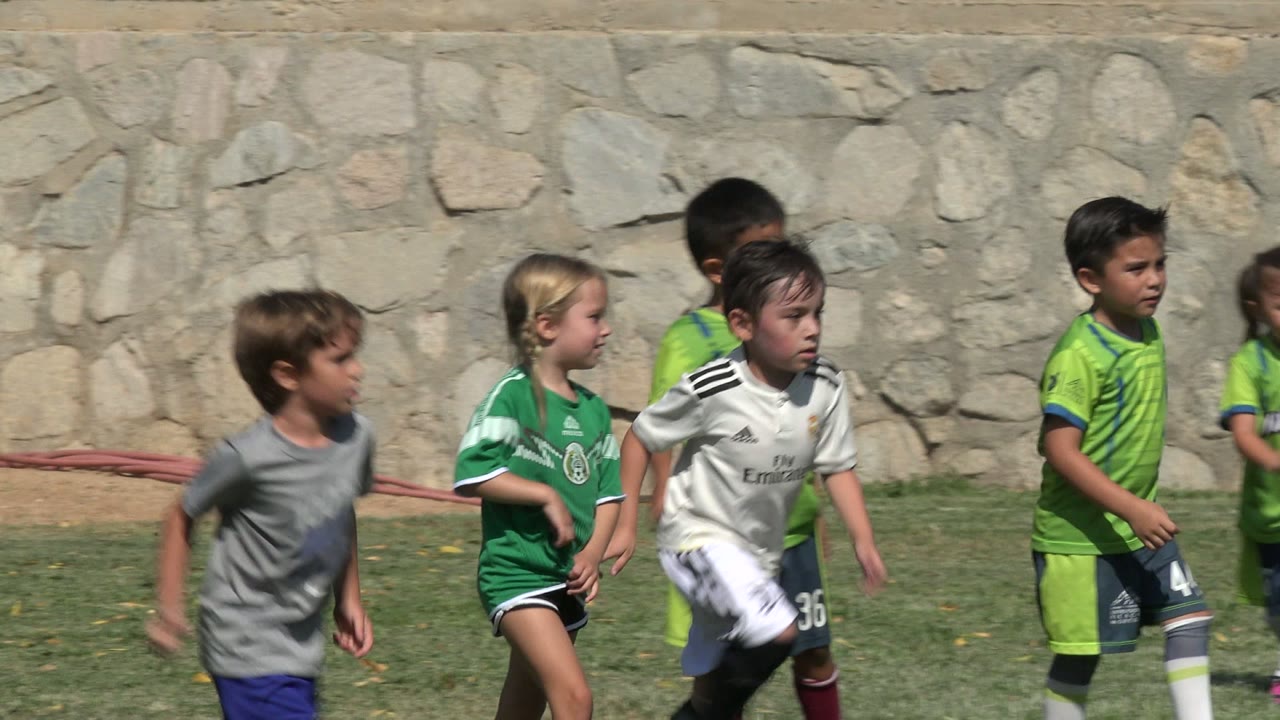 A girl in a green jersey and children in green and white soccer kits move on the grass field in San Jose Del Cabo, Mexico, with bright sun and a stone wall behind them.