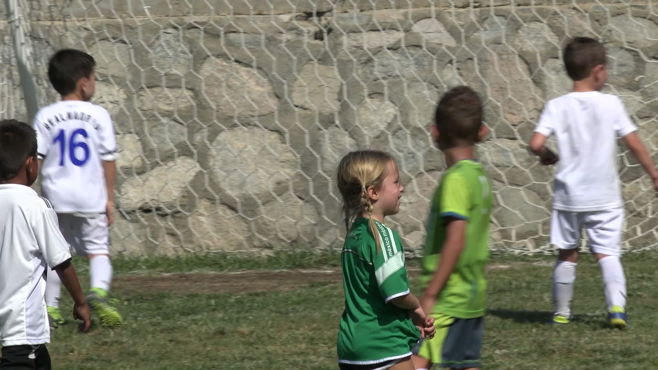 Several children in white and green soccer jerseys gather in front of the goal net in San Jose Del Cabo, Mexico, while a child in a neon goalkeeper shirt stands near the post.