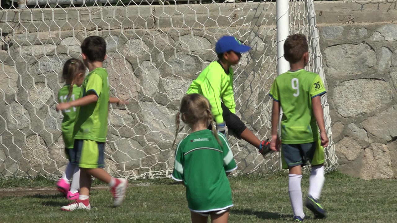 A young goalkeeper in a neon green shirt and blue cap stands beside the goal net while other children in green uniforms move across the grass in San Jose del Cabo, Mexico.