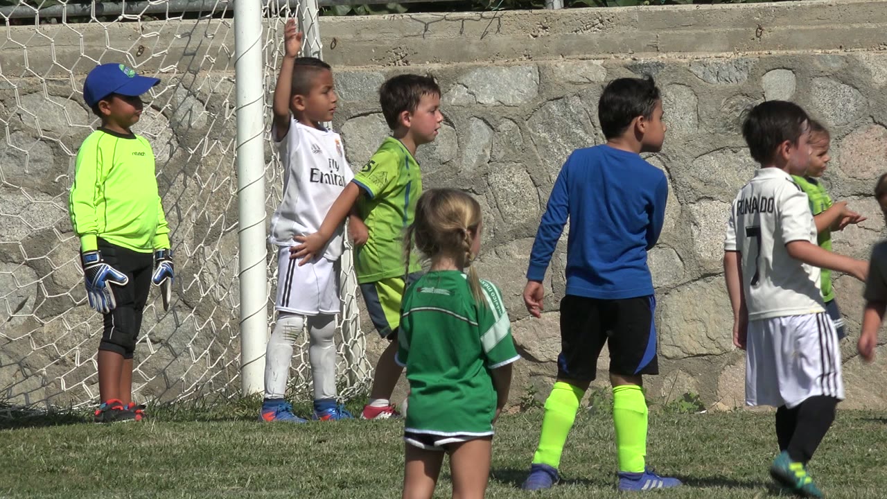 A child in a white soccer kit raises an arm near the goal while other children stand close together on the grass in San Jose del Cabo, Mexico.