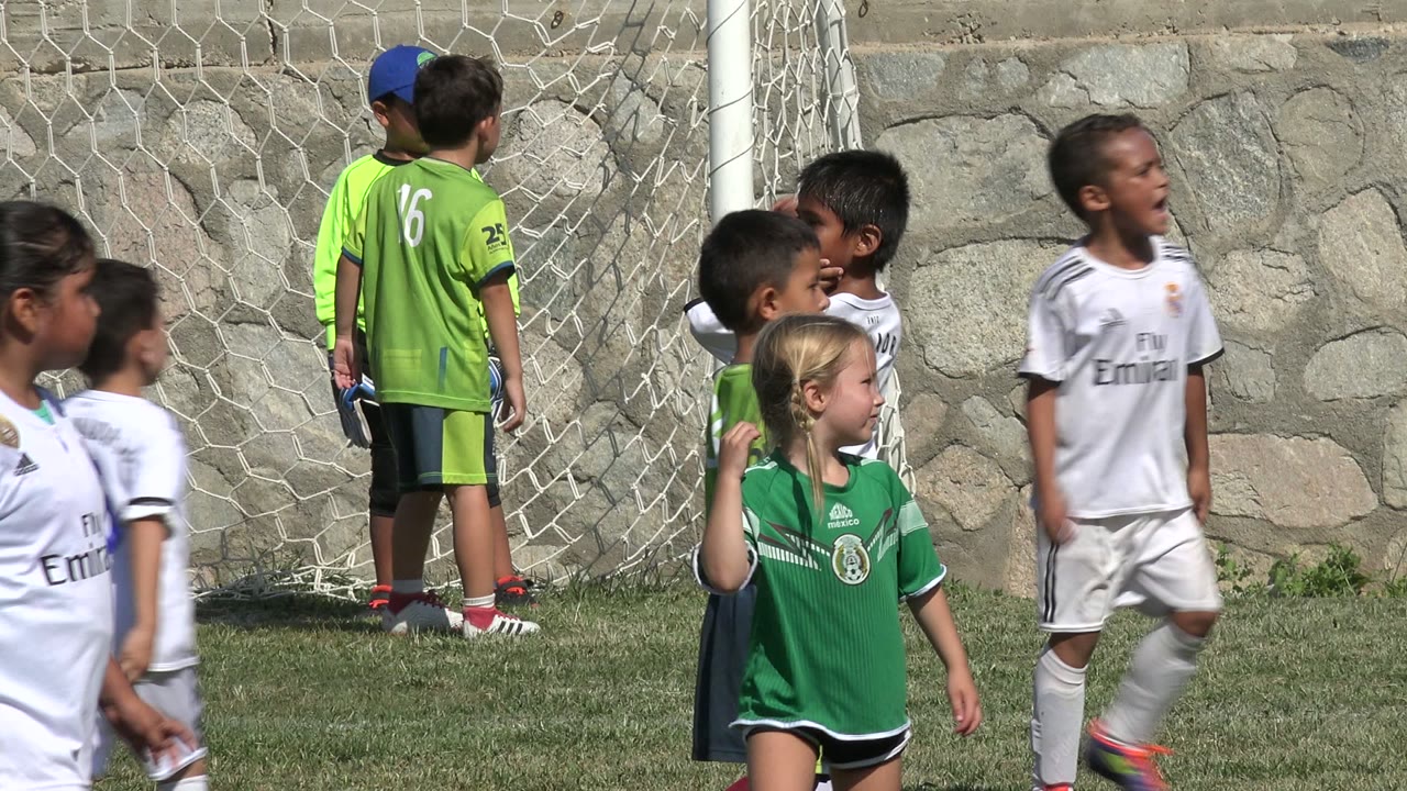 Youth soccer players in mixed green and white uniforms stand and walk beside the goal net, with the stone wall visible behind the field in San Jose Del Cabo, Mexico.