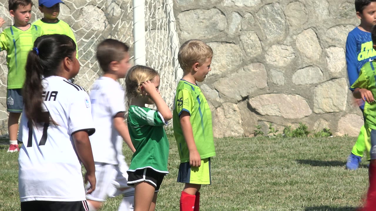 Children in green and white soccer uniforms stand near a goal net and stone wall during soccer practice in San Jose del Cabo, Mexico.