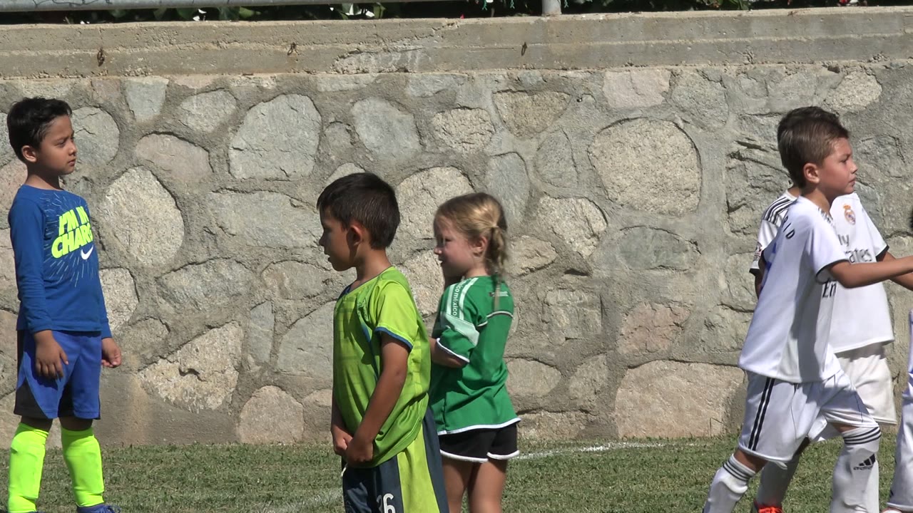 Children in soccer uniforms stand and walk on a grass field near a stone wall.