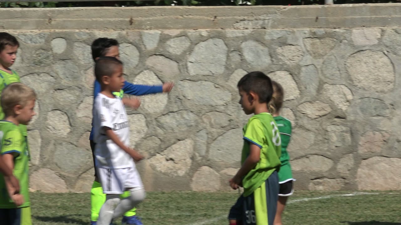 Several children in white and green soccer uniforms cross the grass in San Jose del Cabo, Mexico, with the stone wall behind them and sunlight on their faces.