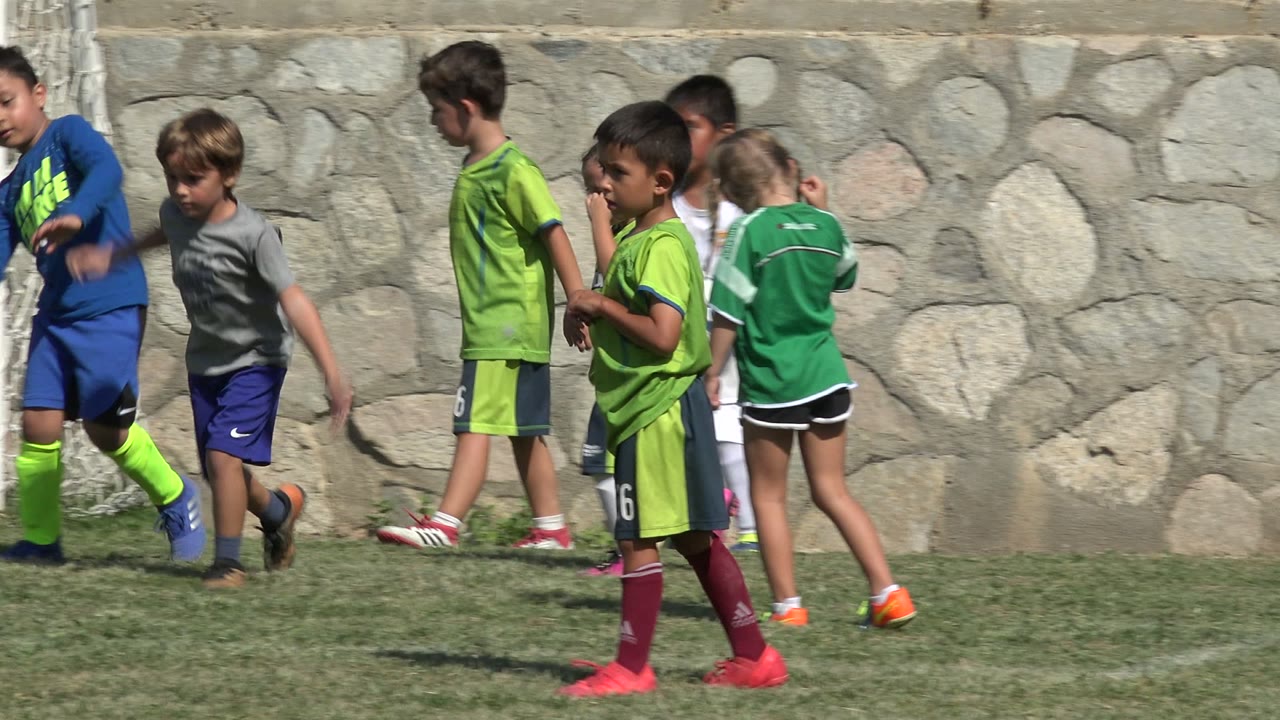 Children in green and blue soccer uniforms move across a grass field in San Jose del Cabo, Mexico, with a stone wall behind them.