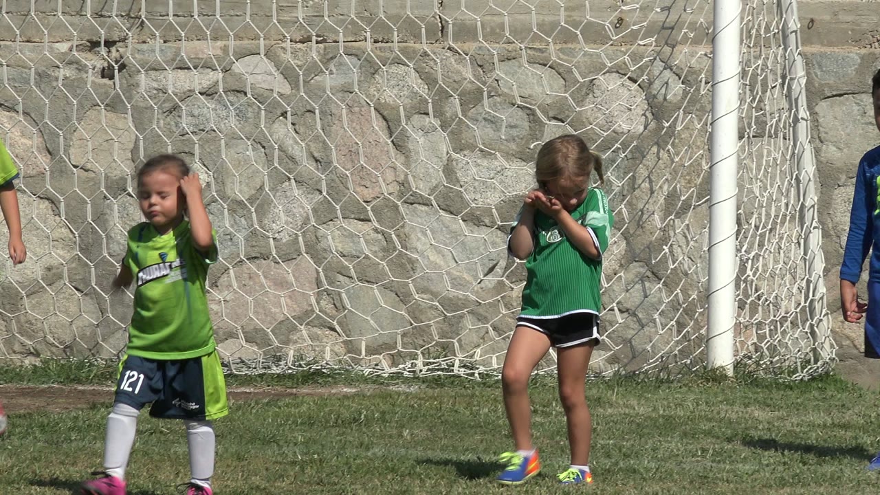A boy in blue and a child in green stand in bright sun beside a goal net, with a stone wall filling the background in San Jose Del Cabo, Mexico.