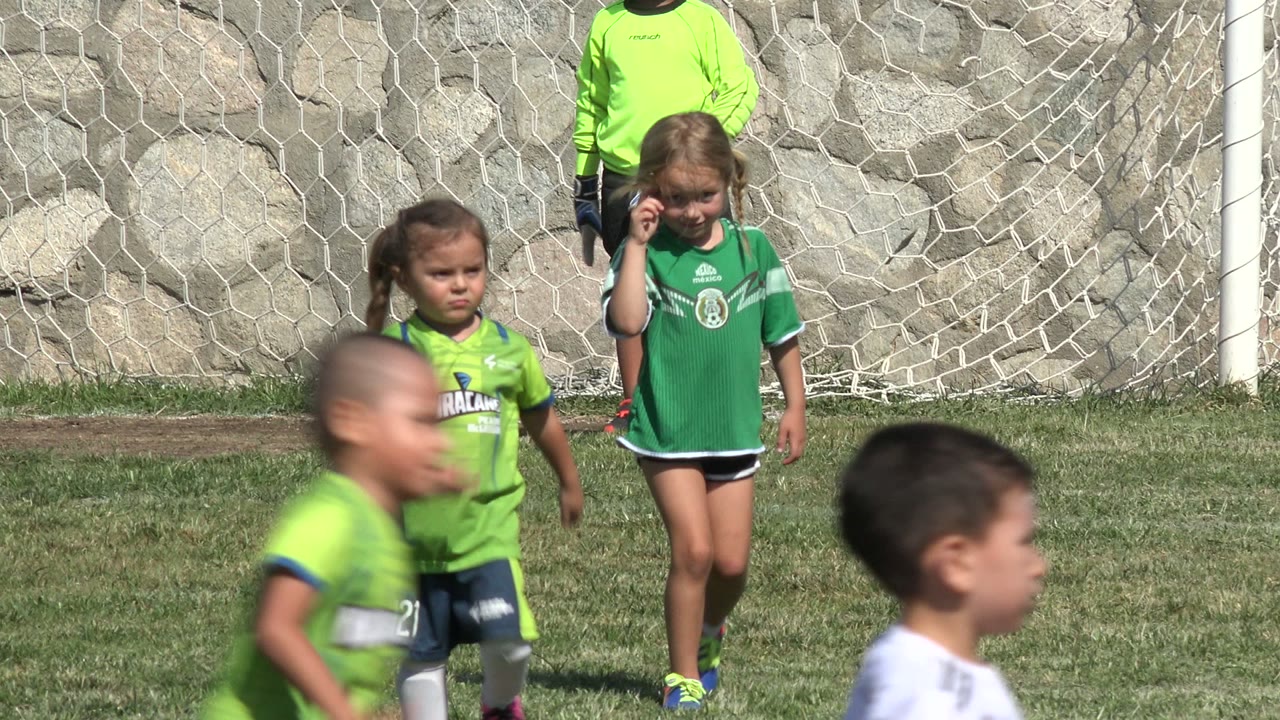 Two young girls in green soccer uniforms stand in front of the goal net in San Jose del Cabo, Mexico, while children in white and green uniforms move across the foreground.